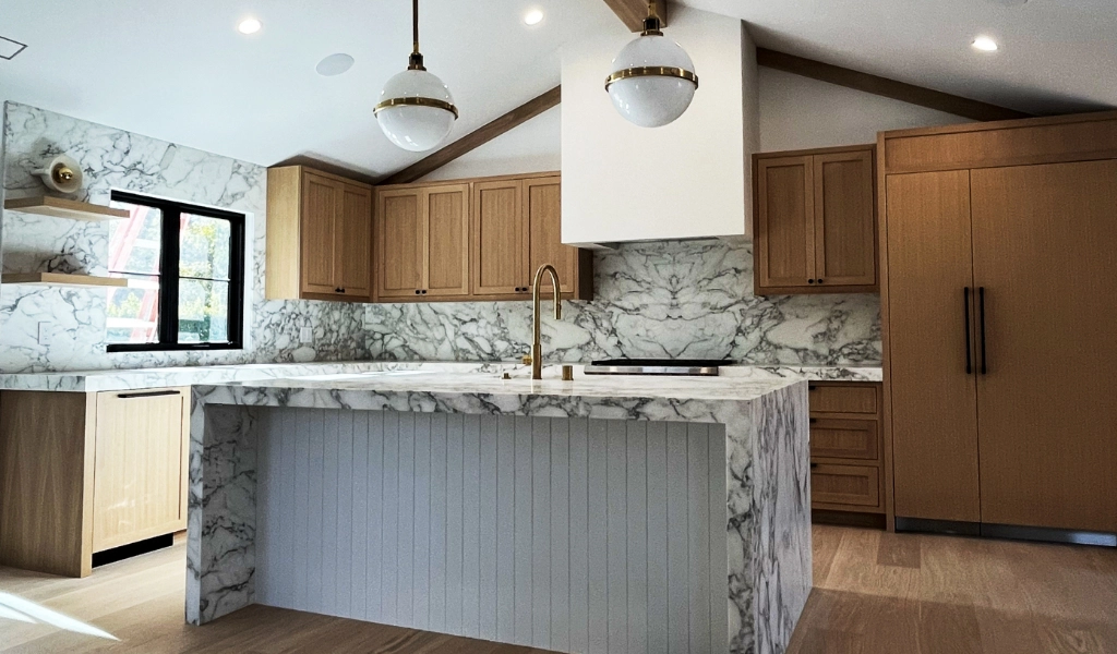 A contemporary kitchen featuring a marble island, marble countertops, wooden cabinets, two ceiling-mounted globe lights, and a built-in refrigerator.