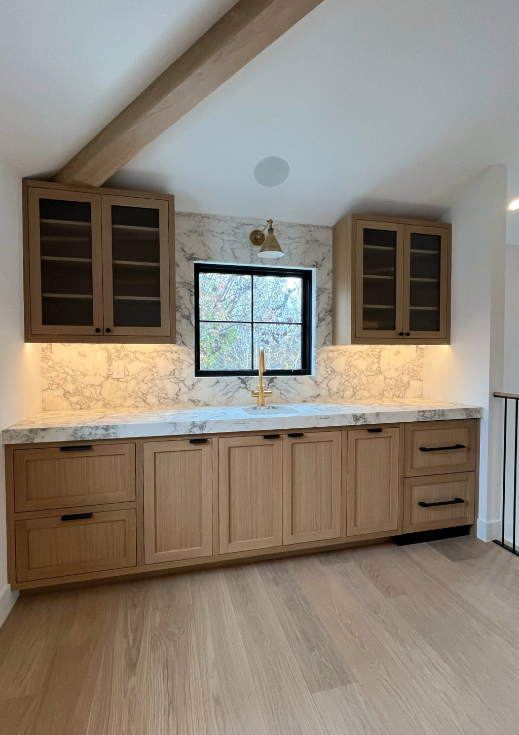 Modern kitchen with light wood cabinets, marble countertops, and backsplash, featuring a central window framed in black, open shelves, and a brass faucet under warm recessed lighting.