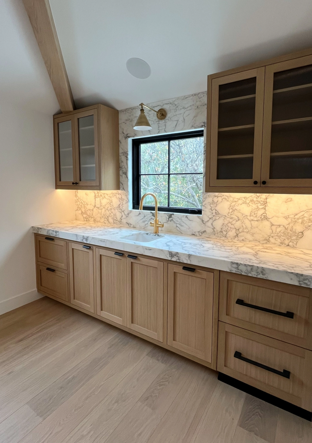 Kitchen with light wooden cabinets, marble countertops, black-framed window, and a gold faucet. Upper cabinets have glass doors; a wall sconce provides lighting above the sink area.