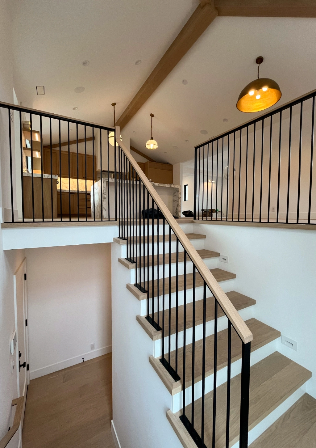 Interior view of a modern two-story home with a staircase featuring wooden steps and black metal railings; kitchen area is visible upstairs.