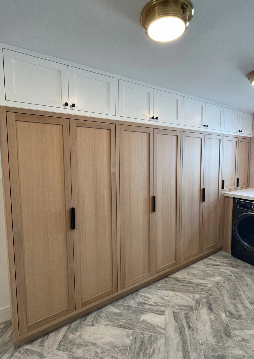 A laundry room features light wood-finished cabinetry with black handles, white upper cabinets, a washer and dryer, and stone tile flooring.