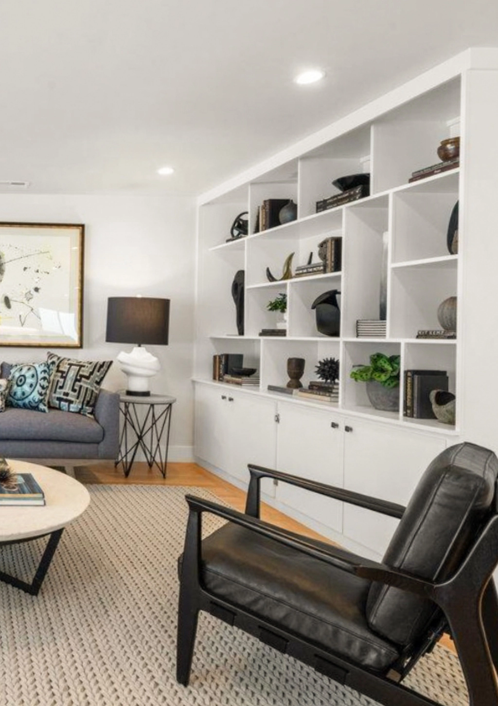 A modern living room with a white bookshelf, black leather armchair, round coffee table, blue couch, and table lamp. Neutral tones and decorative items create a minimalist aesthetic.