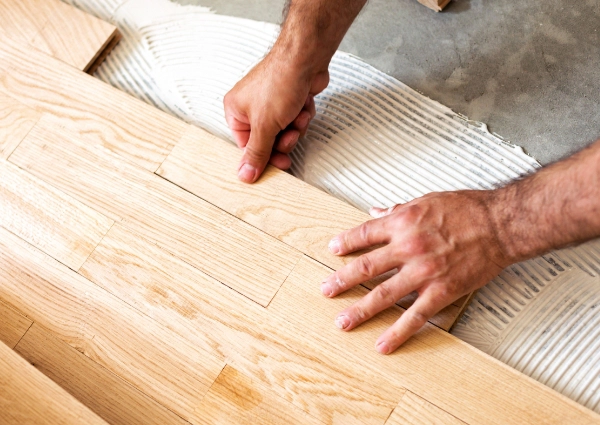 Hands placing light-colored wooden flooring planks over adhesive, a key step in home remodeling.