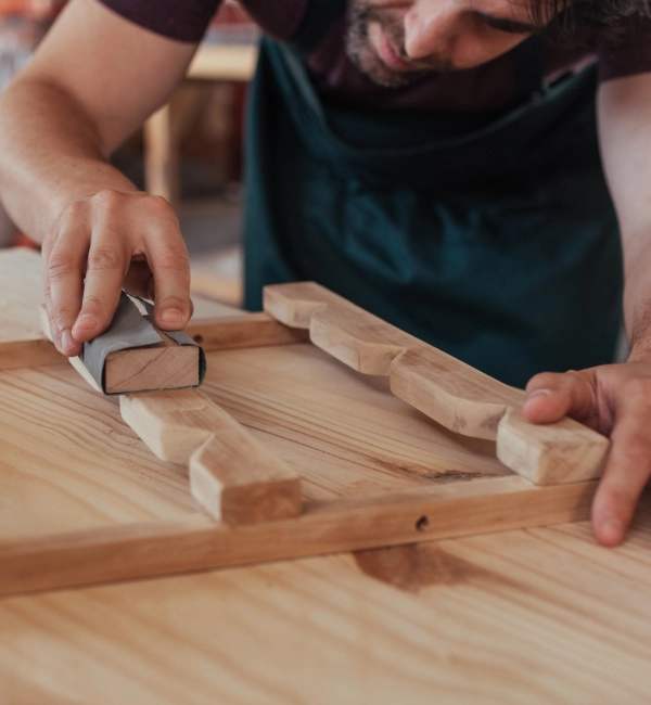 A person in a dark apron sands the wooden frame of a furniture piece with a sanding block.