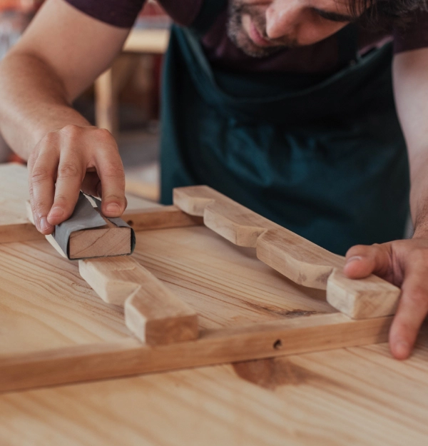 A person wearing an apron sands the wooden surface of a furniture piece, working intently in a workshop at Ciarrai Construction.