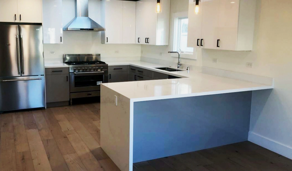 Modern kitchen with stainless steel appliances, white cabinets, grey lower cabinets, a large island with white countertops, a built-in oven and stove, and wooden flooring.