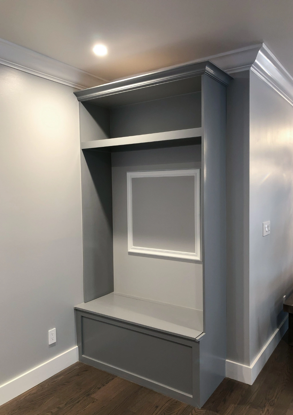 A custom bathroom features a built-in gray wall unit with a bench, an upper shelf, and a framed section on the back, situated in a corner of the room with recessed lighting and hardwood flooring.
