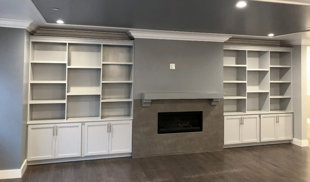 A room with a fireplace centered between two built-in shelving units and cabinets. The cabinets and shelves are painted white, complemented by the dark hardwood floors, creating a cohesive design that echoes the elegance of a custom bathroom.
