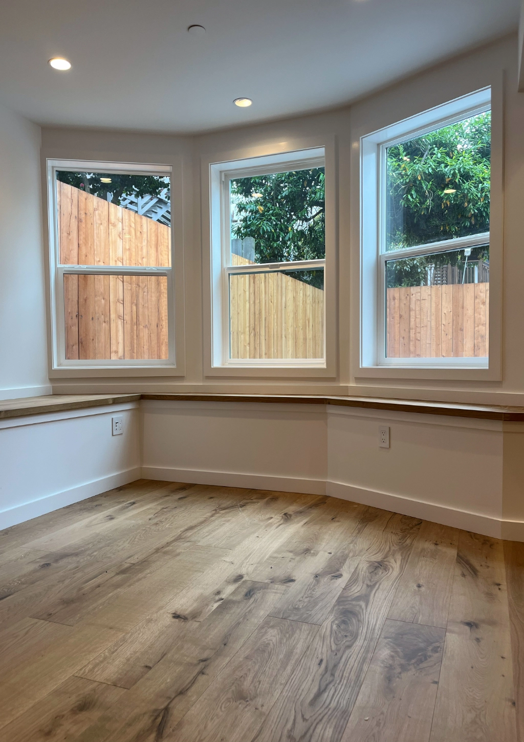 A small room with three large windows, light wood flooring, recessed lighting, and a view of a wooden fence and green foliage outside could easily be transformed into a custom bathroom.