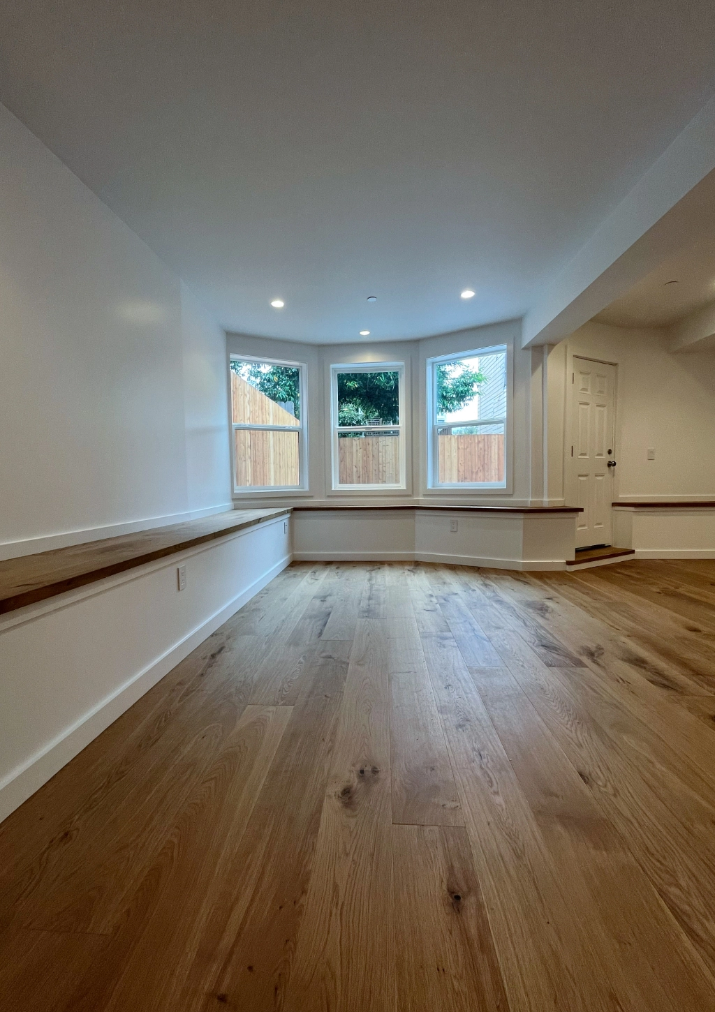 A basement room with wooden flooring, white walls, three large windows, and a single door to the right. Sunlight is streaming in from the windows, illuminating the wooden bench along the wall. Adjacent to this inviting space is a custom bathroom for added convenience.