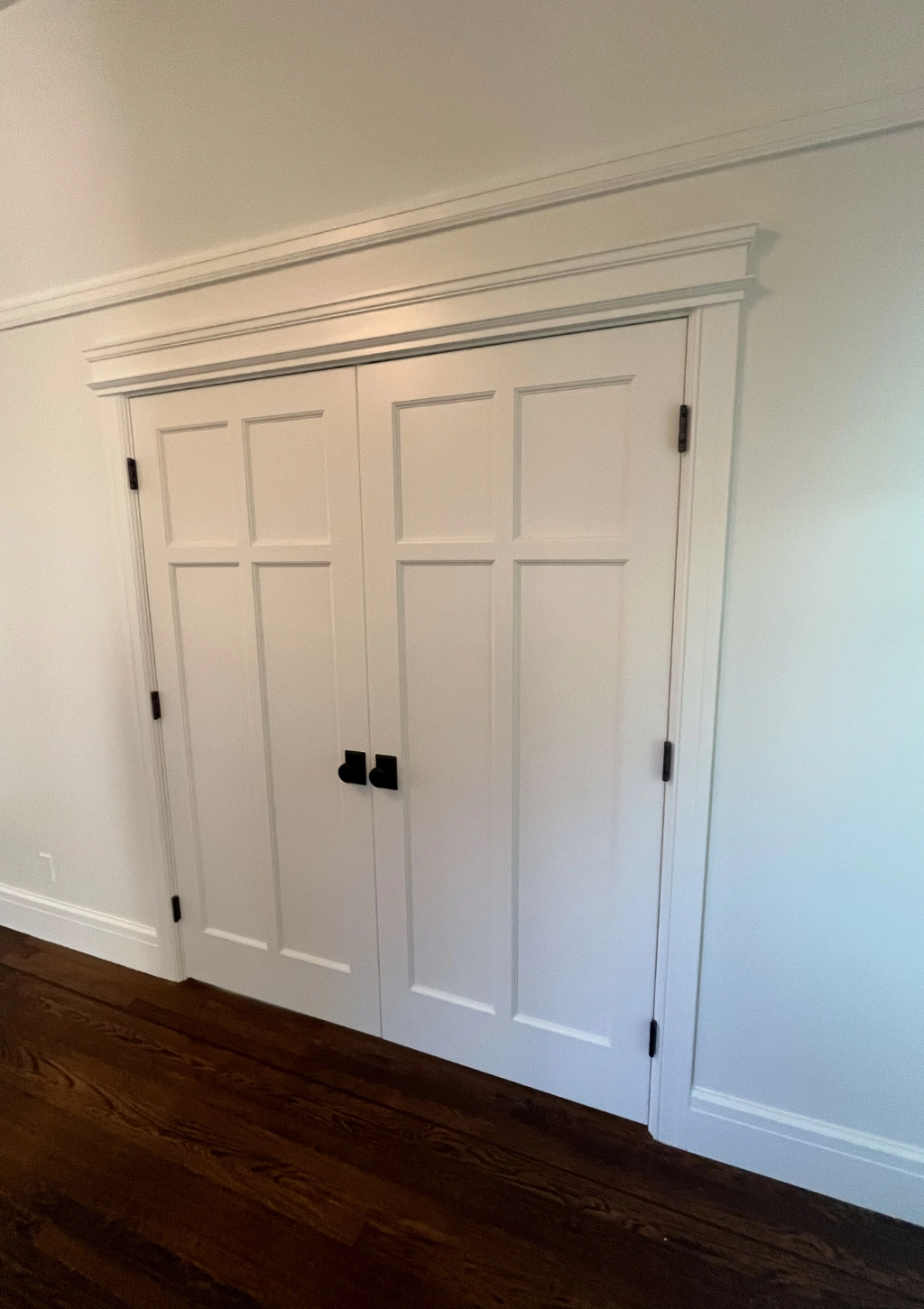A set of closed white double doors with black handles, framed by a white door trim, is situated against a white wall. Dark brown hardwood flooring is visible at the bottom, leading into what could be a custom bathroom beyond the elegantly designed entrance.