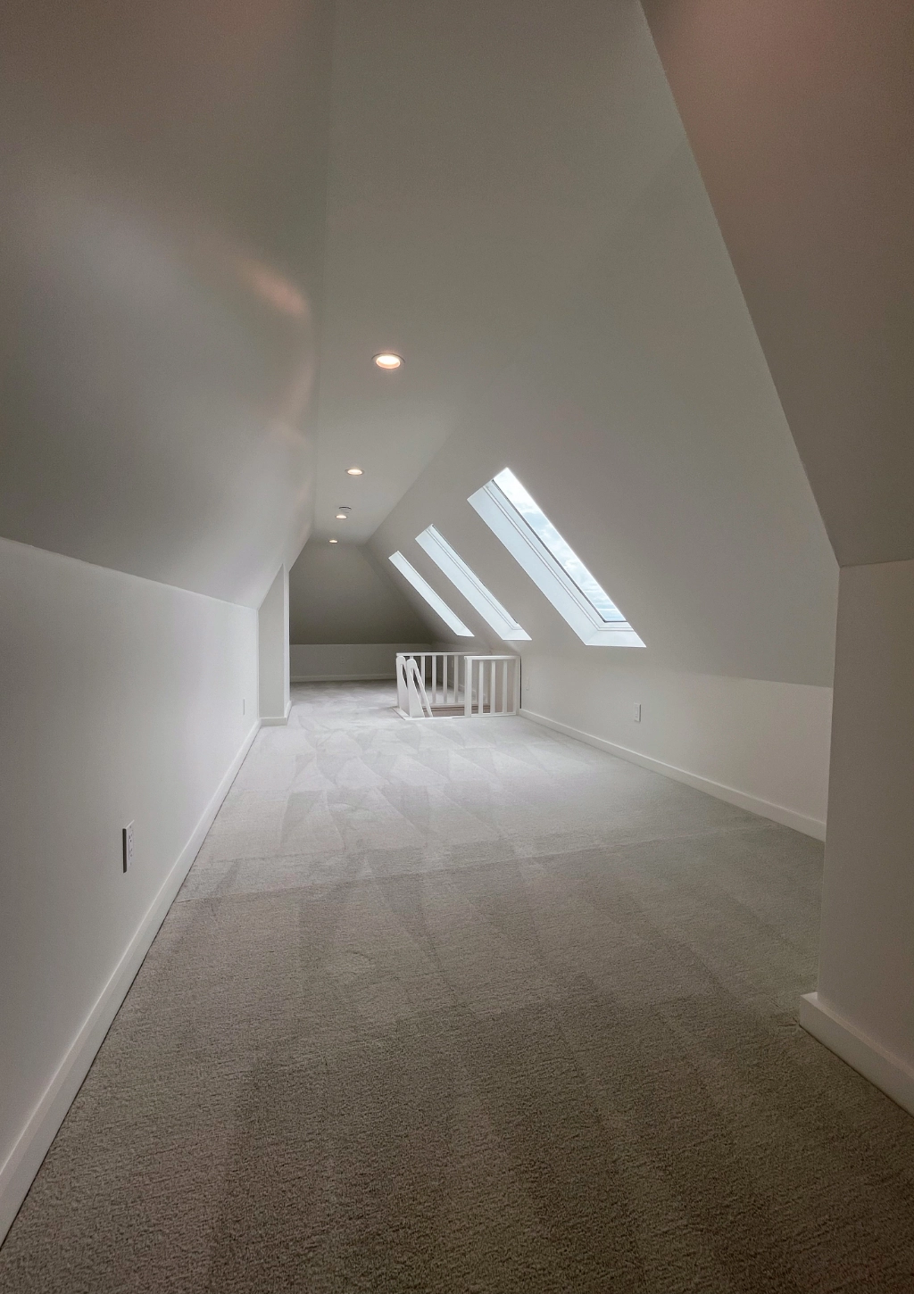 A narrow, carpeted attic room with sloped ceilings, skylights, and a staircase leading to the lower level. Spaces are painted white and include a custom bathroom for added convenience.