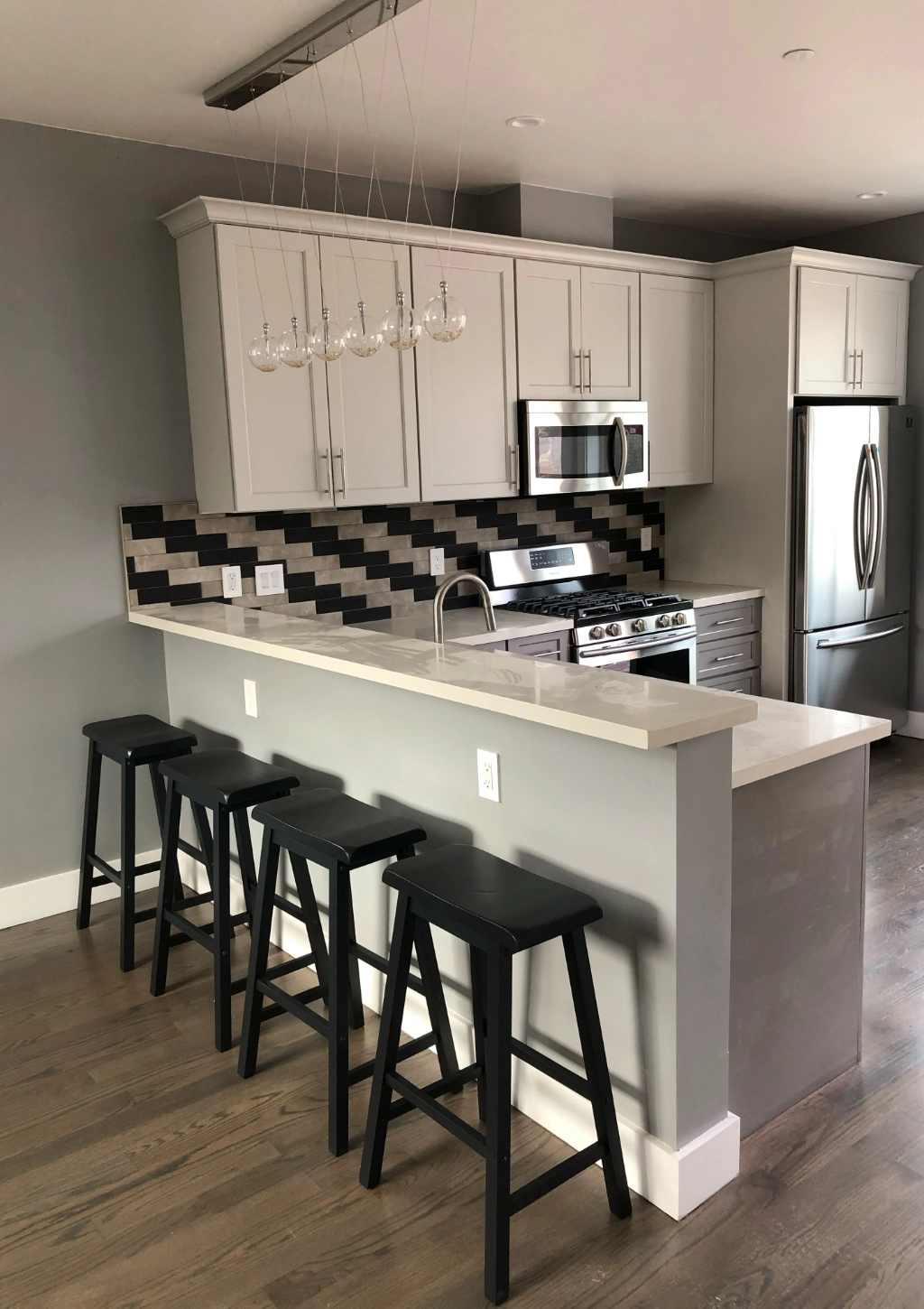 Modern kitchen with grey cabinets, stainless steel appliances, and a white countertop. Four black stools are aligned at the counter. The backsplash features a black and grey geometric pattern, complementing the custom bathroom design in this stylish home.