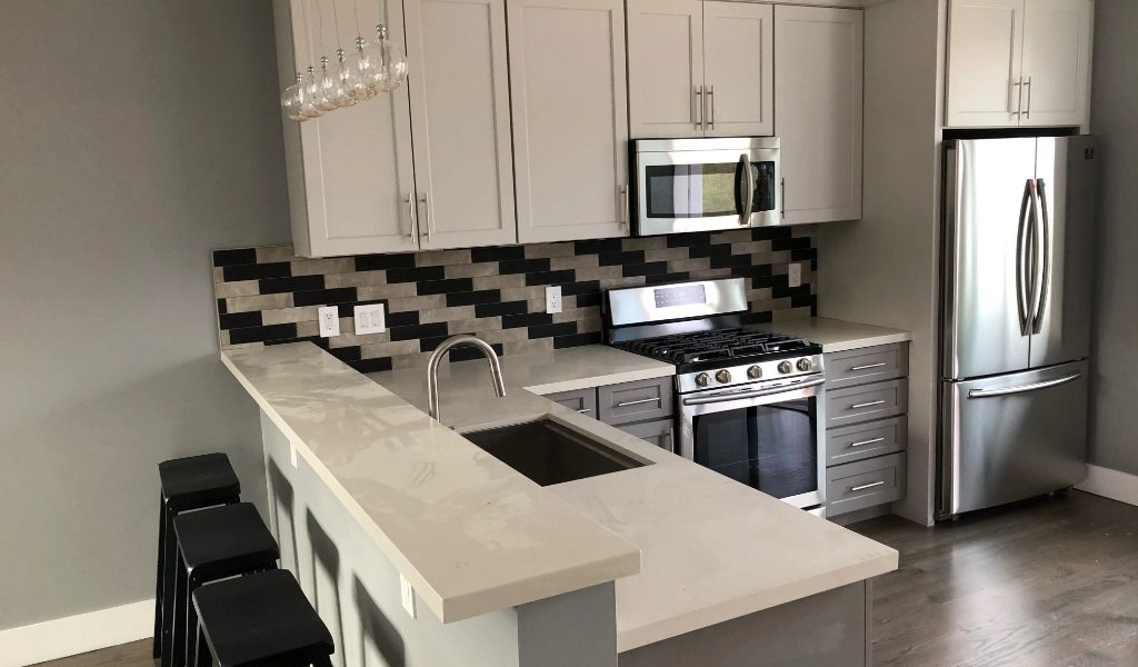 Modern kitchen with white cabinets, stainless steel appliances including a refrigerator, microwave, and oven, and a black and white backsplash. An island with a sink and bar stools is in the foreground, seamlessly connected to a custom bathroom for added convenience.