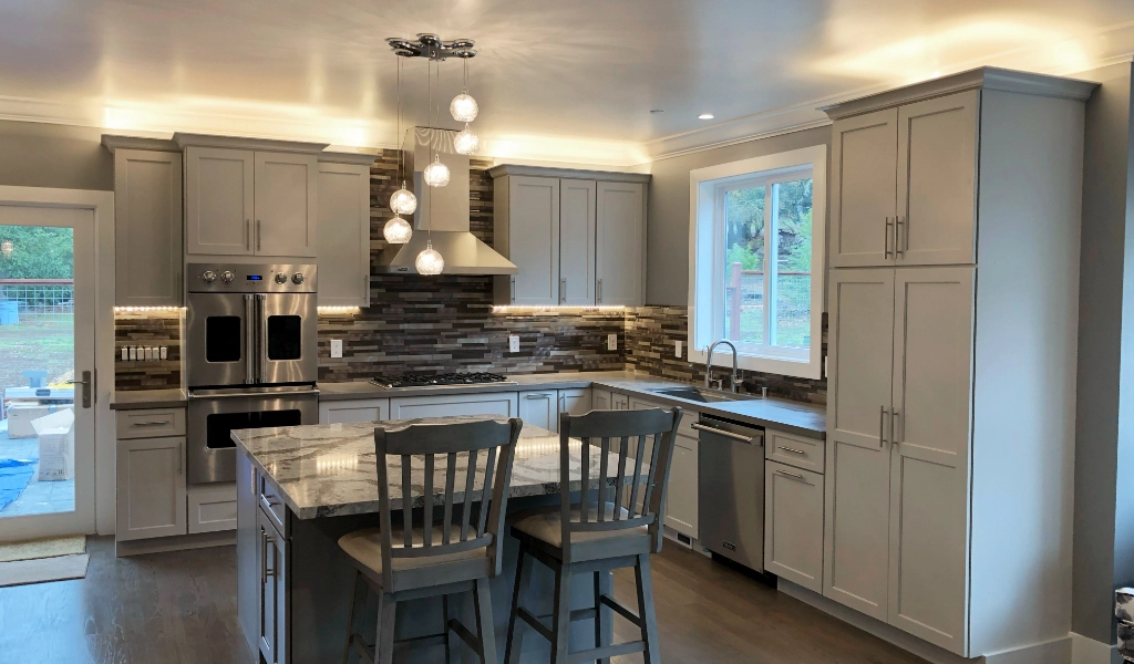 Modern kitchen with two chairs at an island, stainless steel appliances, white cabinets, marble countertops, and a backsplash with gray and brown tiles. Pendant lights hang above the island. This home features not only a stylish kitchen but also a custom bathroom to complete the luxurious feel.