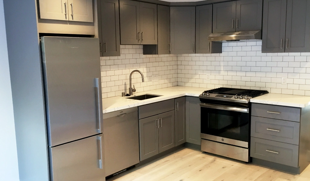 Modern kitchen with gray cabinets, stainless steel appliances including a refrigerator and oven, white subway tile backsplash, and light wood flooring seamlessly connected to a custom bathroom featuring sleek fixtures.