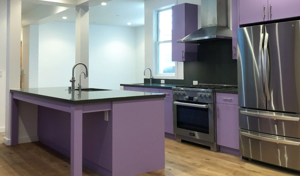A modern kitchen features purple cabinets, a large island with a sink, stainless steel appliances, and hardwood floors. The design seamlessly connects to a custom bathroom, adding an extra touch of luxury and coherence to the living space.
