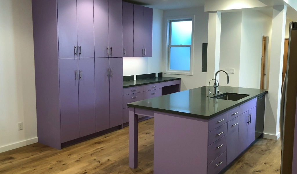 A modern kitchen with light purple cabinets, black countertops, and wooden flooring. A window is above the sink and recessed lighting brightens the space, complementing the sleek design found in a custom bathroom.