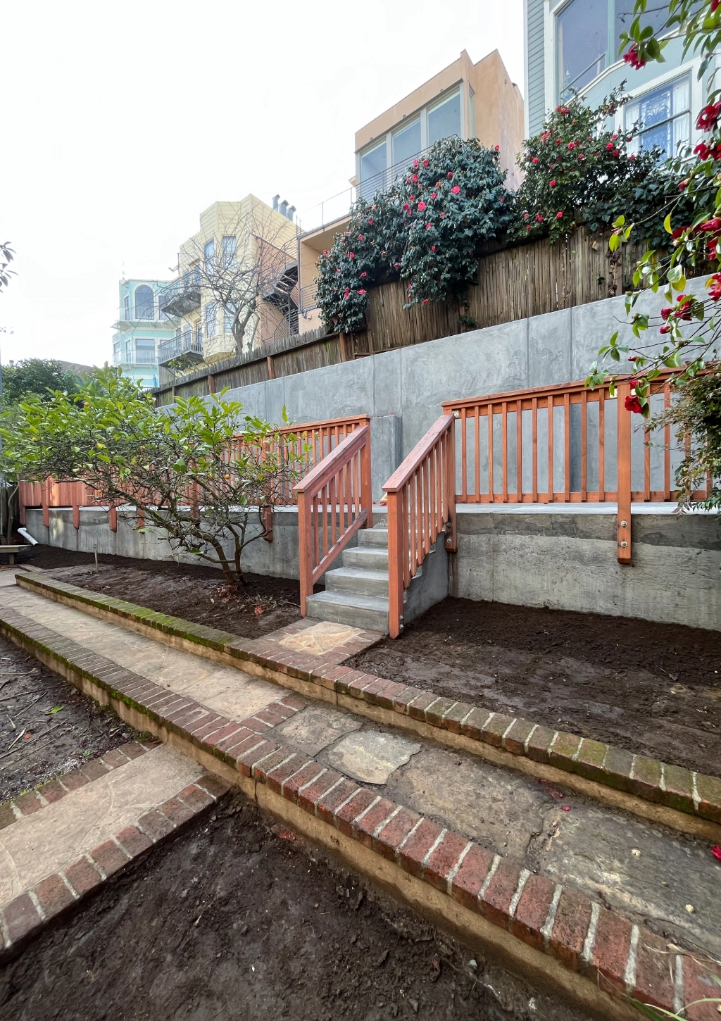 A two-tiered garden area with concrete walls and stairs, wooden railings, and surrounding plants. Brick pathways are visible on both levels, creating a serene outdoor space akin to the tranquility of a custom bathroom.