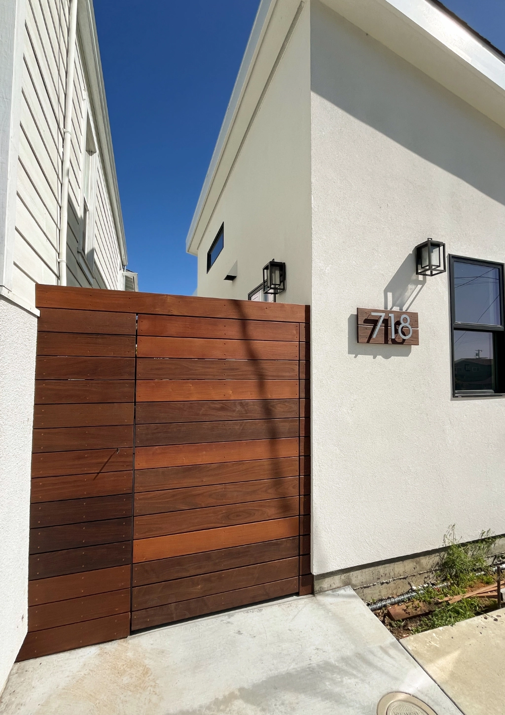 Modern white building exterior with a wooden gate and the number "718" displayed on the wall. Two black wall-mounted lights are above the number, hinting at an elegant interior that might just feature a custom bathroom. The sky is clear and blue.