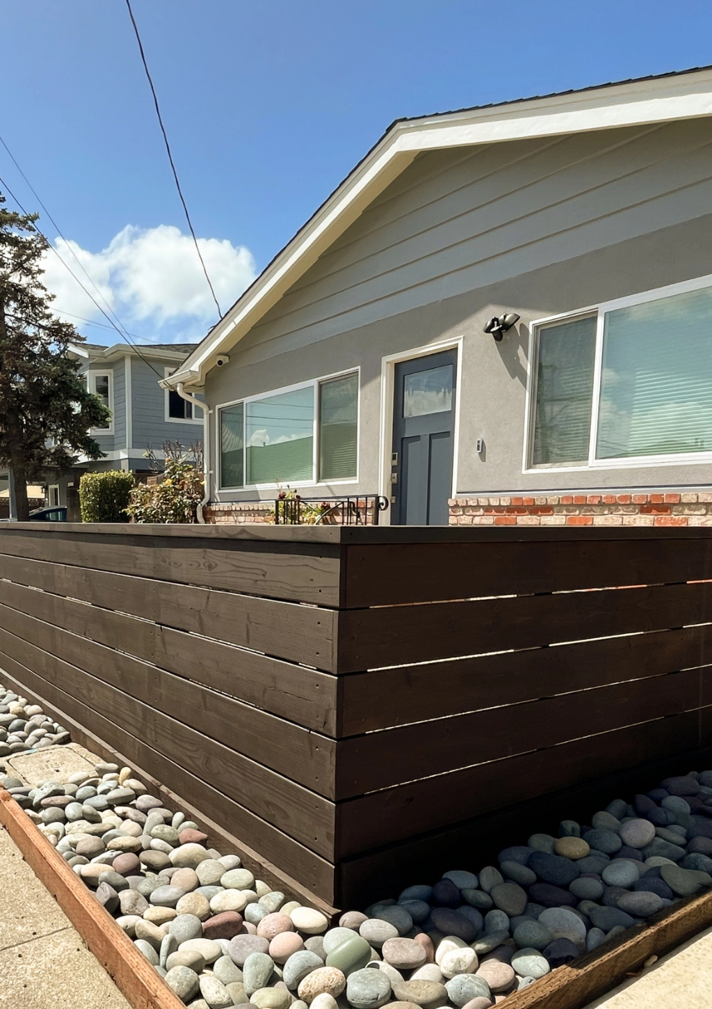 A light grey house with a dark wooden fence and rock landscaping in the front yard. The house, featuring large windows and a grey door, also boasts a custom bathroom that adds a touch of luxury to its well-designed aesthetic.