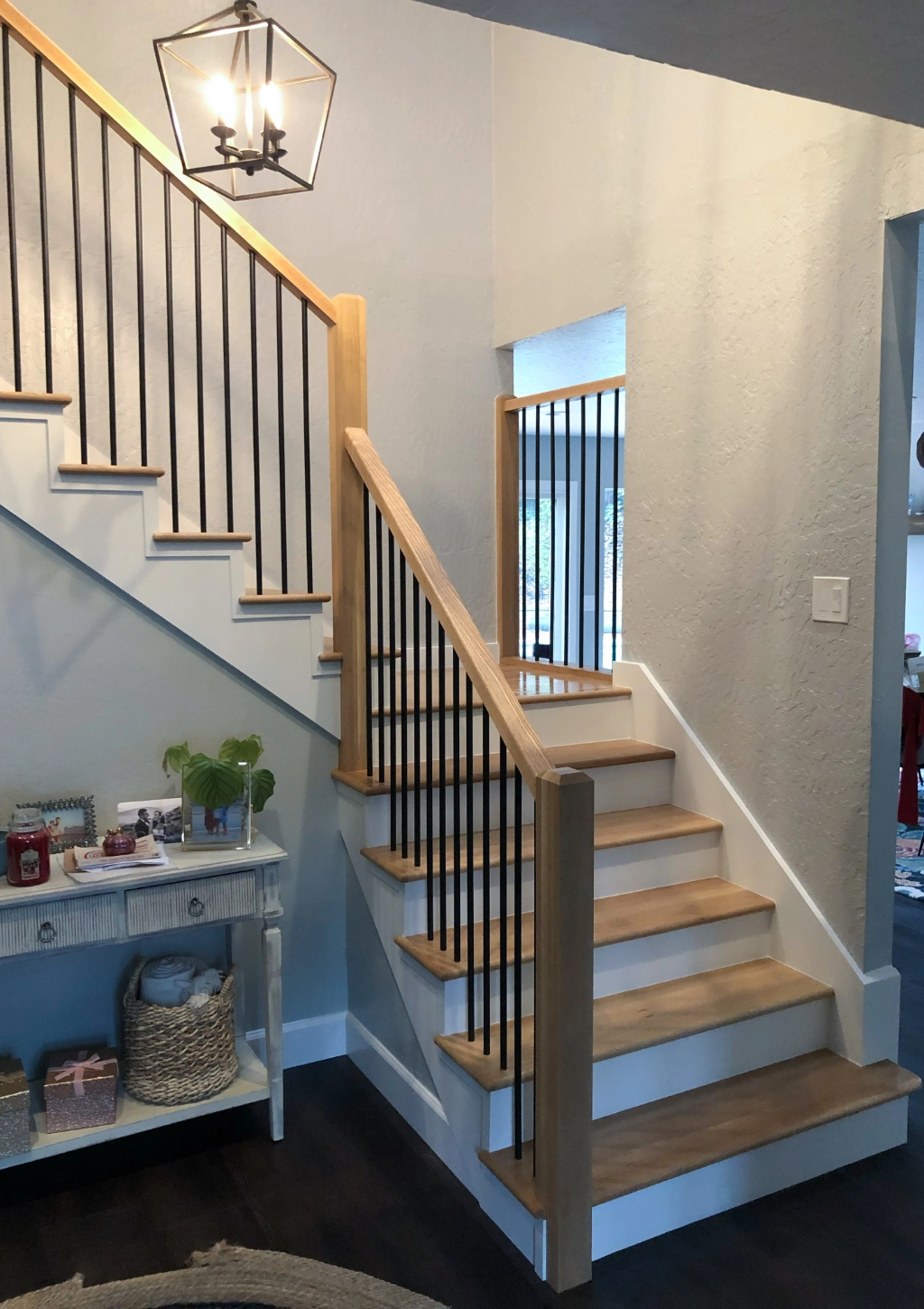 A wooden staircase with black metal balusters and a lantern-style light fixture is shown, beside a small white side table topped with various decorative items, accentuating the home's elegant design, which seamlessly extends into the custom bathroom.