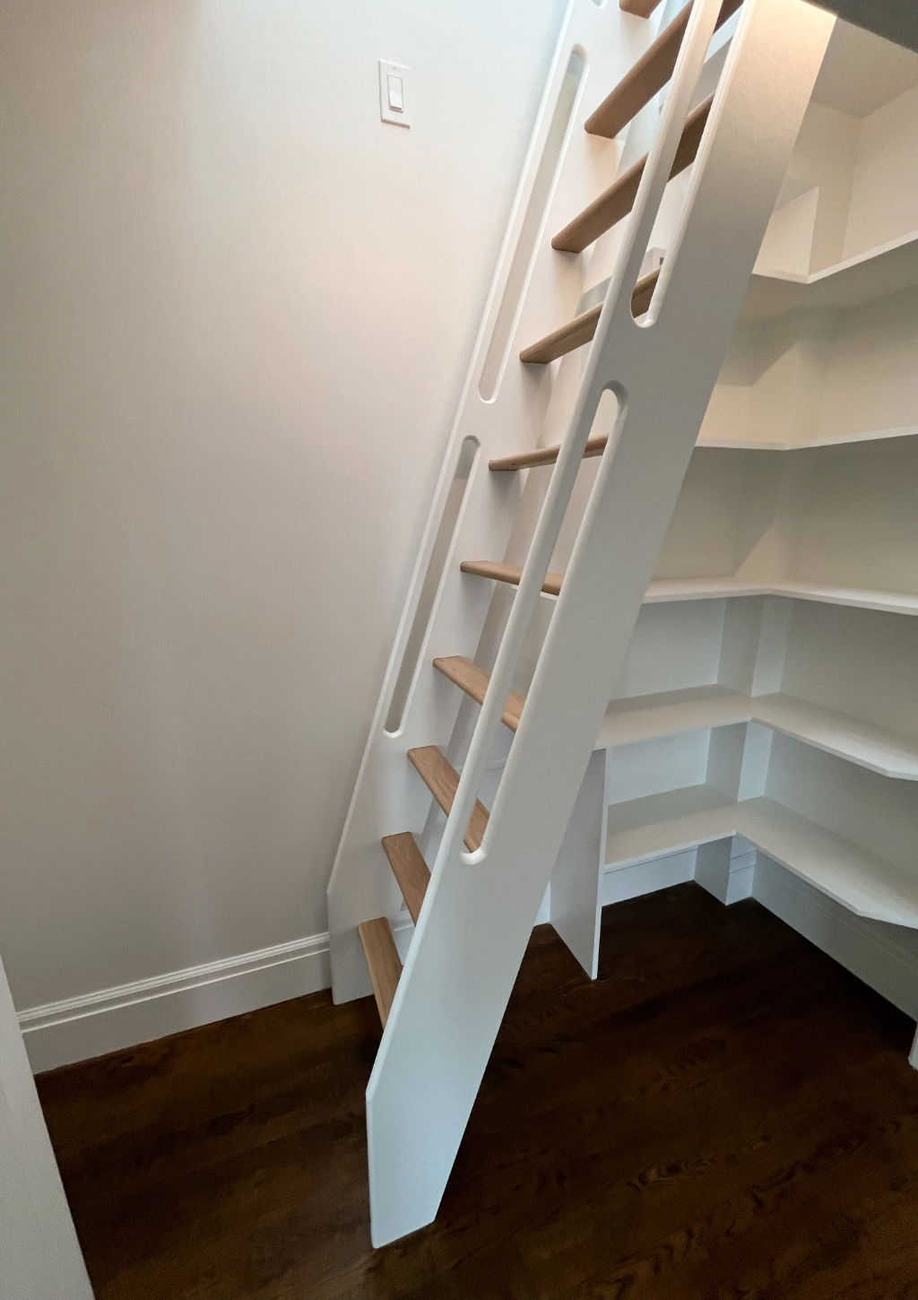 A white, wooden ladder staircase with light brown steps leads to an upper level next to corner shelves on a dark hardwood floor, seamlessly blending into the custom bathroom's elegance.