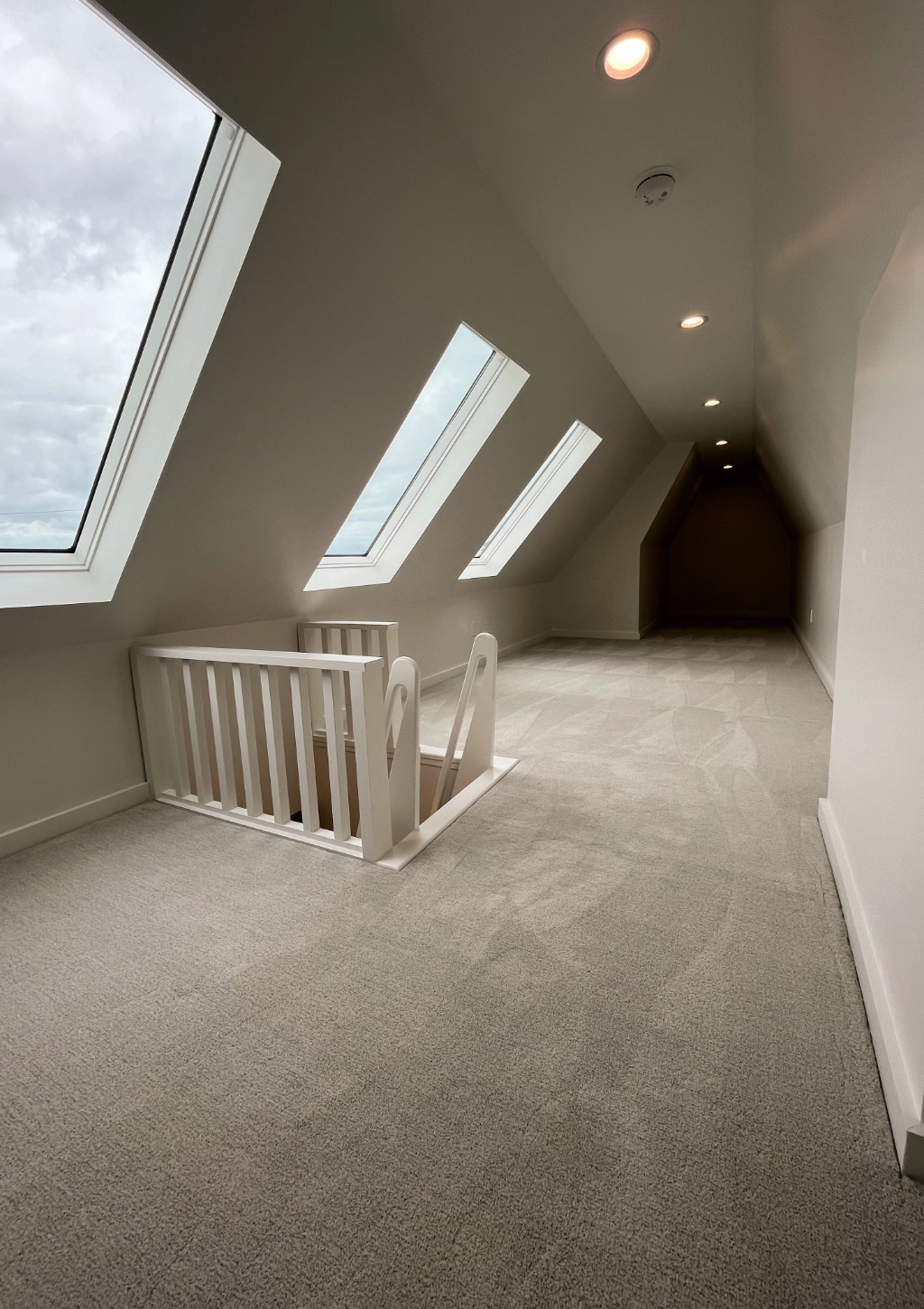 A long, narrow, carpeted attic space with skylights, recessed lighting, and a white wooden railing around a stairwell opening leads to a custom bathroom.