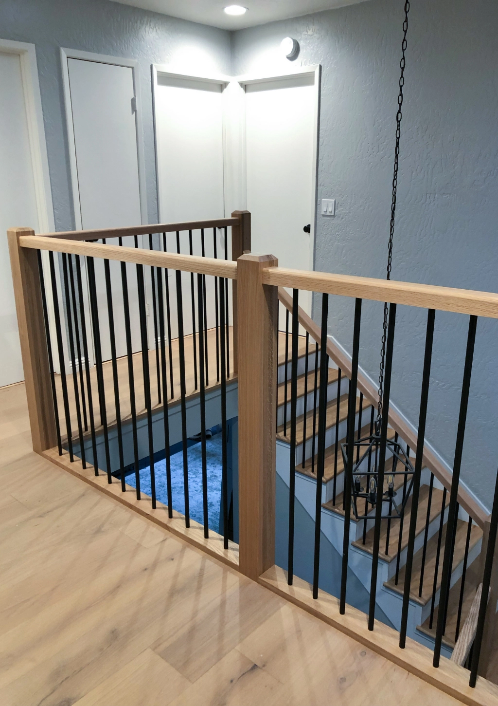 Wooden staircase with black metal balusters leading downstairs, featuring light gray walls and two closed white doors at the landing. A ceiling light fixture and wooden flooring are visible, showcasing a path towards a custom bathroom nearby.